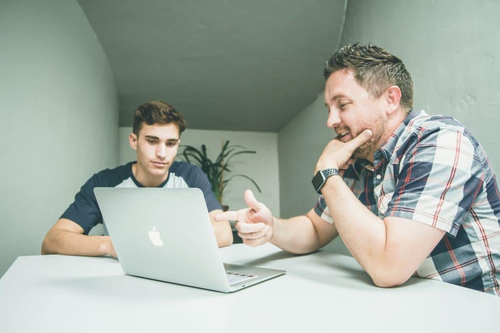 Man showing a young man something on a laptop sitting at a desk.